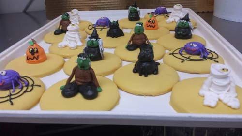 A tray of decorated cookies featuring Halloween-themed characters like monsters, pumpkins, and spiders on yellow bases.