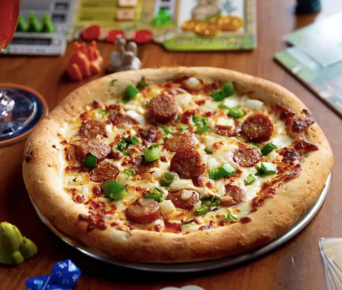 A delicious pizza topped with sausage, green onions, and cheese, surrounded by board game pieces on a wooden table.