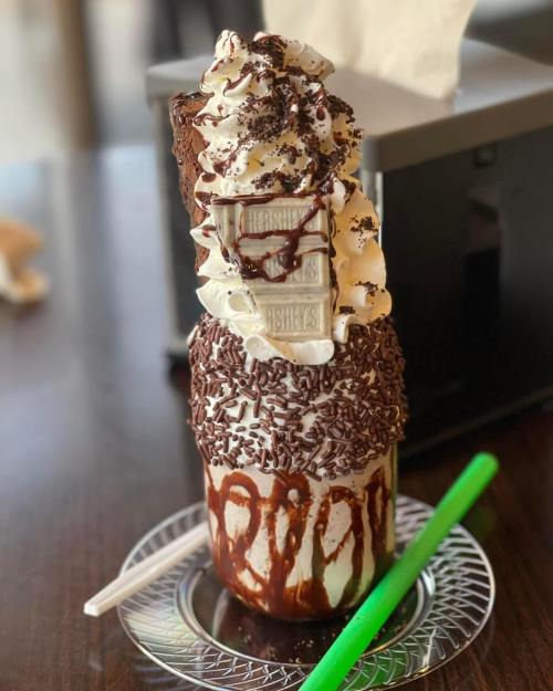 A tall milkshake topped with whipped cream, chocolate sprinkles, and a Hershey's chocolate bar, served on a plate with a straw.