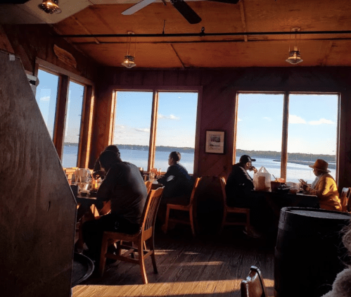 Interior of a restaurant with diners at wooden tables, large windows showing a view of the water outside.