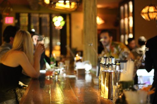 A dimly lit bar scene with patrons at the counter, drinks on the bar, and a bartender in the background.