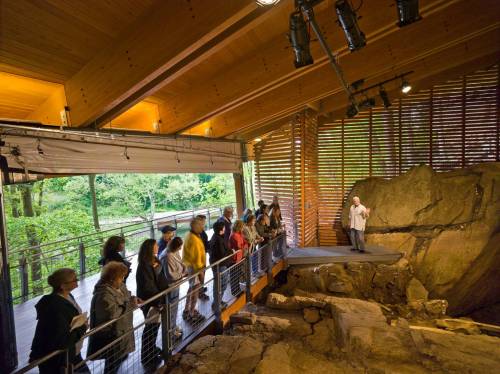 A group of people listens to a speaker in a modern exhibit space showcasing archaeological findings.