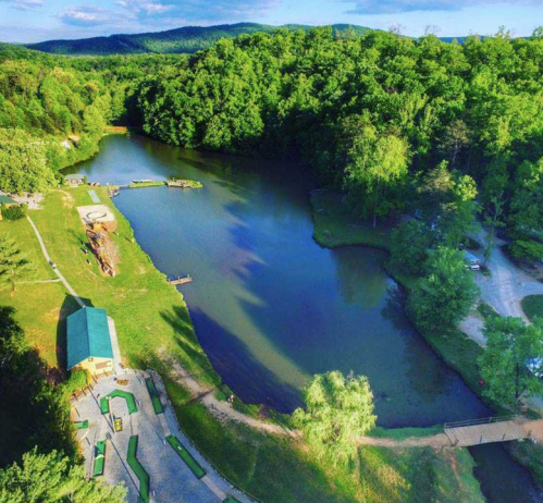 Aerial view of a serene lake surrounded by lush greenery and a small dock, with a path and picnic area nearby.