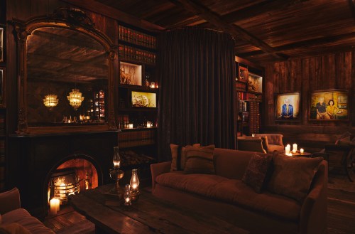 Cozy, dimly lit room with a fireplace, plush sofas, bookshelves, and artwork, creating an inviting atmosphere.