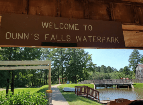 Sign reading "Welcome to Dunn's Falls Waterpark" with a view of green trees and a wooden deck in the background.