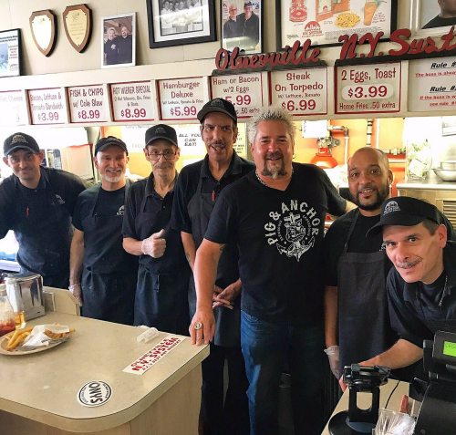 A group of seven men in black aprons stand behind a counter in a diner, smiling at the camera.