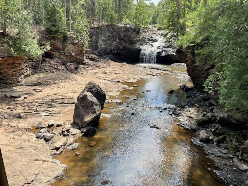 A serene landscape featuring a rocky riverbed, a small waterfall, and lush greenery surrounding the water.