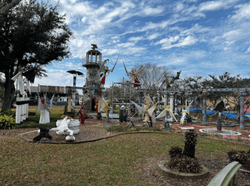 A whimsical outdoor sculpture garden featuring various figures, a lighthouse, and vibrant landscaping under a cloudy sky.