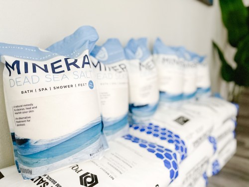 Bags of mineral dead sea salt for bath, spa, shower, and feet, displayed on a shelf with a plant in the background.