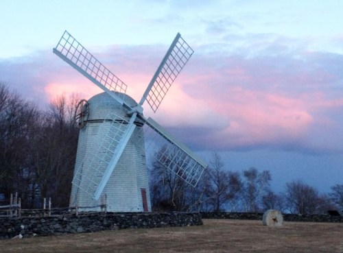 A historic windmill stands against a colorful sunset, surrounded by trees and a stone wall.