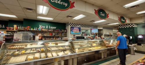 A man stands in front of a deli display case filled with various baked goods and prepared foods in a grocery store.