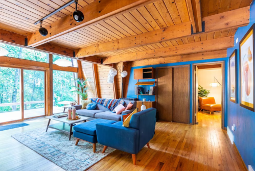 Cozy living room with wooden beams, blue walls, a sofa, and large windows overlooking a forested area.