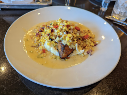 A plate of fried fish topped with corn, peppers, and a creamy sauce, served over a bed of mashed potatoes.