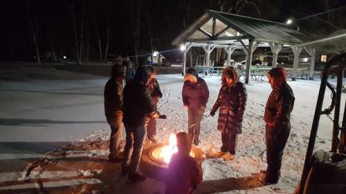 A group of people gathered around a fire pit in a snowy park at night, wearing winter clothing.