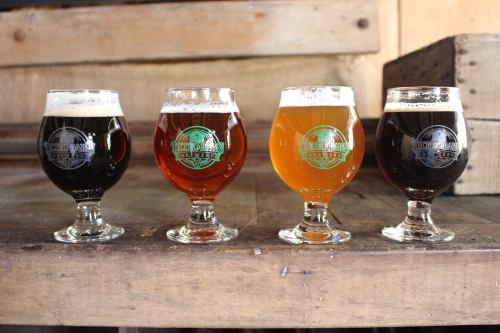 Four glasses of beer in varying colors—dark brown, amber, golden, and black—displayed on a wooden surface.