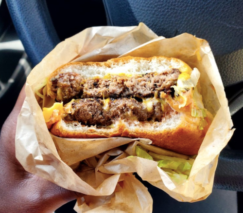 A hand holding a cheeseburger with two patties, lettuce, and cheese, wrapped in brown paper.