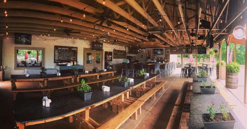 Interior of a rustic restaurant with wooden benches, tables, and a bar area, decorated with plants and string lights.