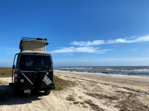 A van with a rooftop tent parked on a sandy beach, with waves and a clear blue sky in the background.