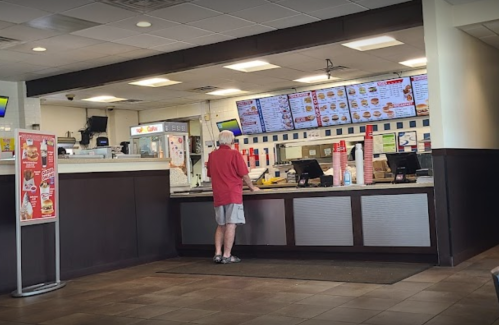 A man in a red shirt stands at the counter of a fast-food restaurant, looking at the menu on the screens above.