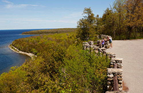 A scenic overlook with stone walls, lush green trees, and a view of a winding coastline and blue water.
