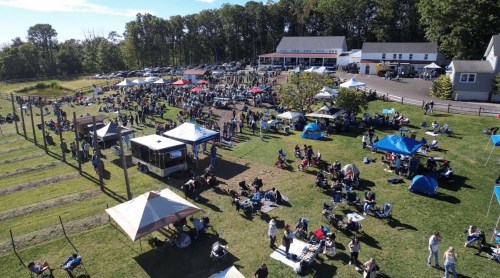 Aerial view of a large outdoor gathering with tents, people, and vehicles in a grassy area surrounded by trees.