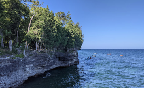 A rocky shoreline with lush trees, overlooking calm blue waters and kayakers in the distance under a clear sky.