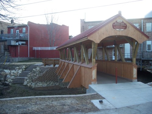 A wooden covered bridge with a red roof, surrounded by buildings and a landscaped area with steps and rocks.