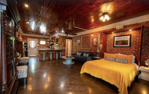 Cozy bedroom with a yellow bedspread, exposed brick walls, wooden ceiling, and a small bar area.