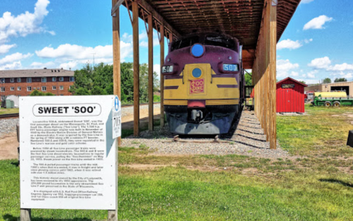 A vintage train displayed under a wooden shelter, with a sign about its history nearby and blue skies above.