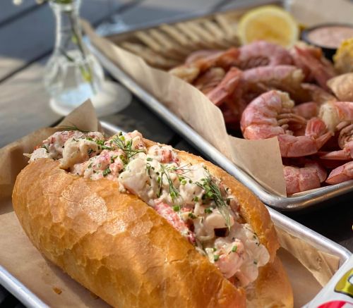 A lobster roll filled with seafood salad, served with shrimp and lemon on a tray.