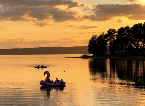 A pink flamingo float on a lake at sunset, with a kayak in the background and trees lining the shore.