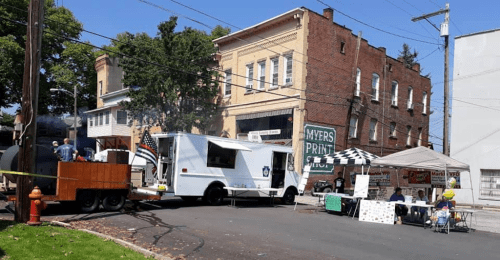 Food truck parked on a street with a tent setup nearby, surrounded by buildings and trees on a sunny day.