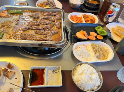 A table set with grilled meat, rice, various side dishes, and drinks, showcasing a Korean BBQ meal.