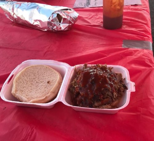 A takeout container with a bun and pulled pork topped with barbecue sauce, resting on a red tablecloth.