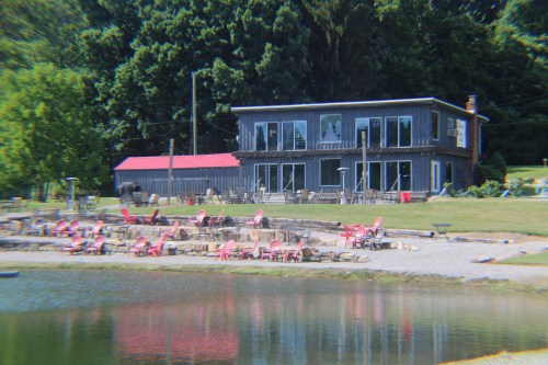 A lakeside view of a modern building with large windows, surrounded by pink chairs and greenery.