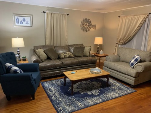 Cozy living room with a brown sofa, armchair, coffee table, and decorative curtains on light walls.