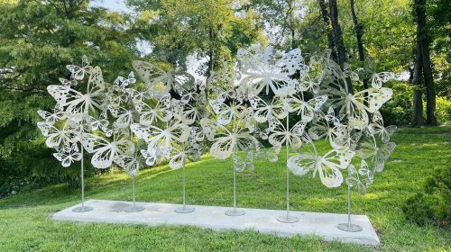 A sculpture of large, metallic butterflies on tall stands, set in a green outdoor area with trees in the background.
