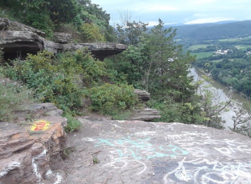 A rocky overlook with graffiti, surrounded by greenery, overlooking a river and valley under a cloudy sky.
