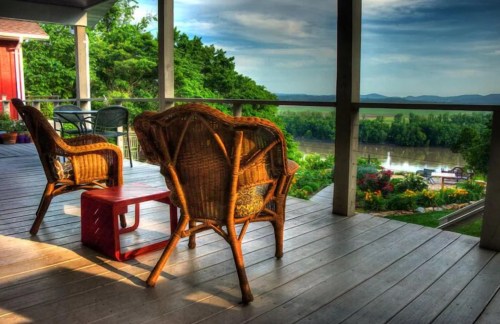 Two wicker chairs on a porch overlook a scenic view of a river and lush greenery under a blue sky.