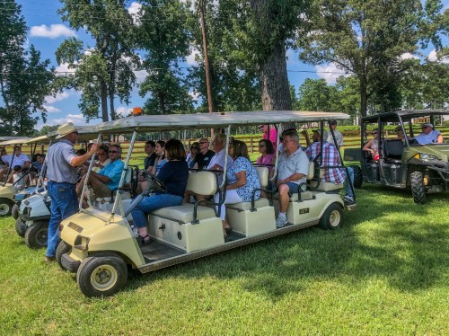 A group of people seated in golf carts, listening to a speaker in a grassy area surrounded by trees.