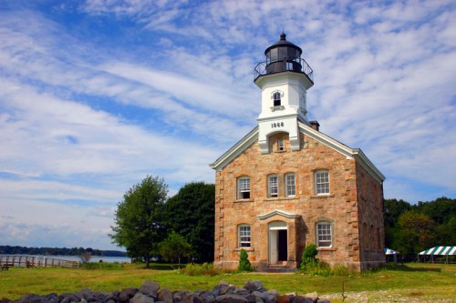 A historic stone lighthouse with a black lantern, surrounded by greenery and a blue sky with scattered clouds.