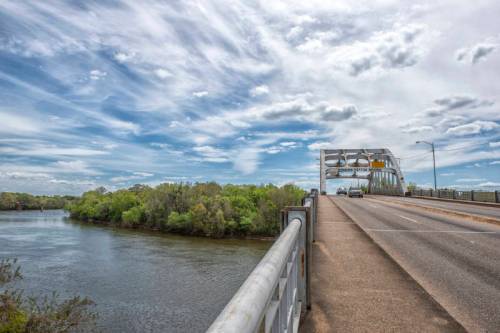 A bridge spans a river under a cloudy sky, with greenery lining the banks and a car driving on the road.