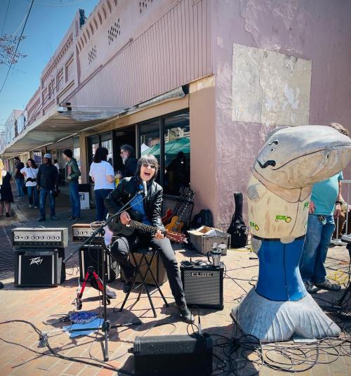 A musician plays guitar on the street next to a large fish statue, with a crowd and shops in the background.