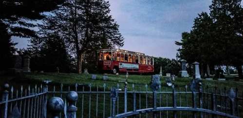 A vintage trolley bus parked on a grassy hill in a cemetery at dusk, surrounded by trees and gravestones.