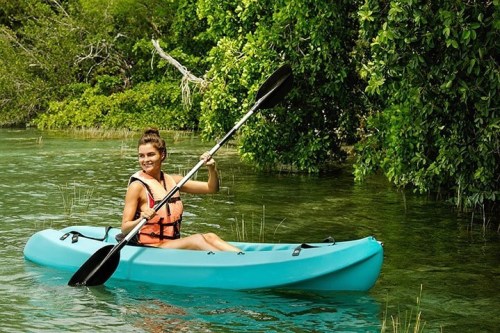 A woman paddles a turquoise kayak through calm, green waters surrounded by lush greenery.
