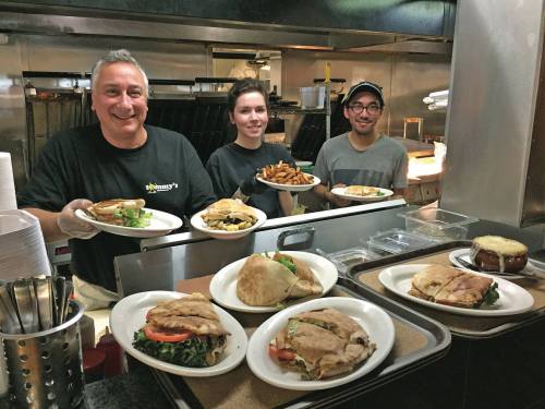 Three restaurant staff members smile while holding plates of food, with various dishes displayed on the counter in front of them.