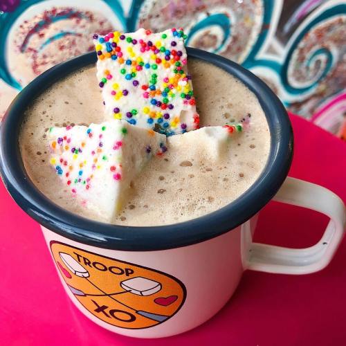 A colorful mug filled with frothy hot chocolate topped with sprinkles and two festive, decorated marshmallows.