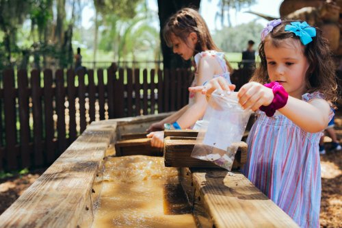 Two children play at a water table, one pouring a bag of items into the flowing water, surrounded by greenery.
