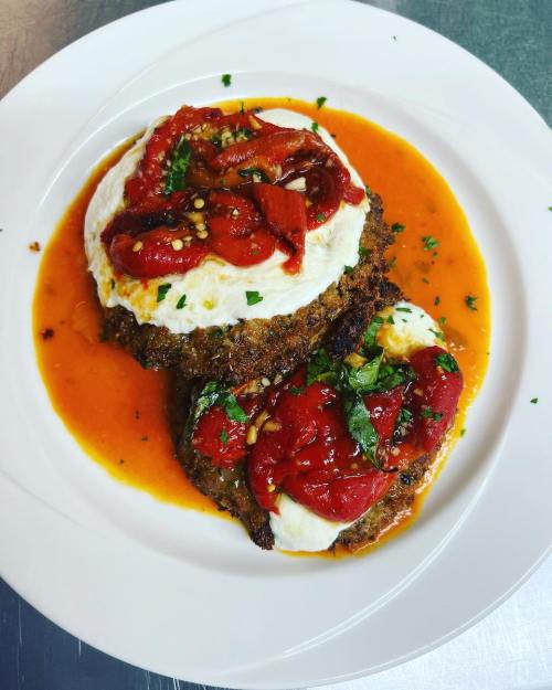 A plate of two crispy patties topped with poached eggs, sun-dried tomatoes, and a rich red sauce, garnished with herbs.