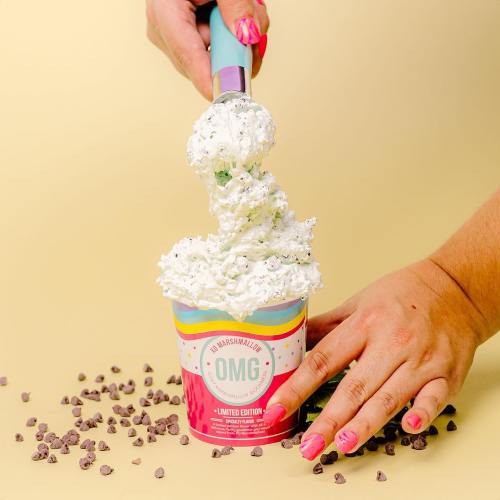 A hand scooping a colorful ice cream topped with whipped cream and chocolate chips from a container on a yellow background.